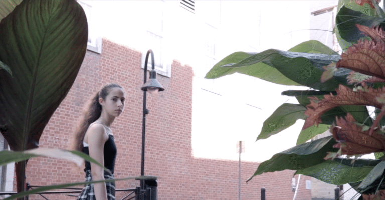 Teen girl near brick building, framed by lush plants.