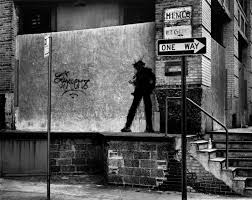 Shadowy figure walking on a ledge in an urban alleyway, next to a "One Way" sign.