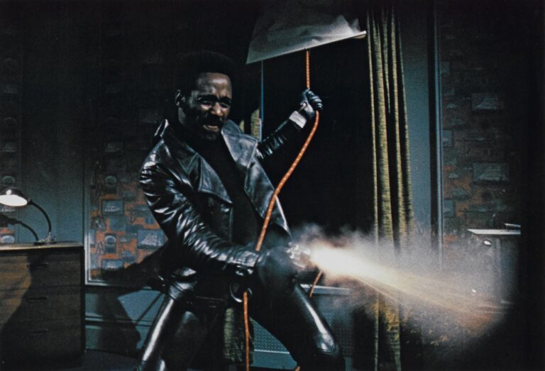 Shaft, in a leather jacket, fires a weapon with sparks flying.