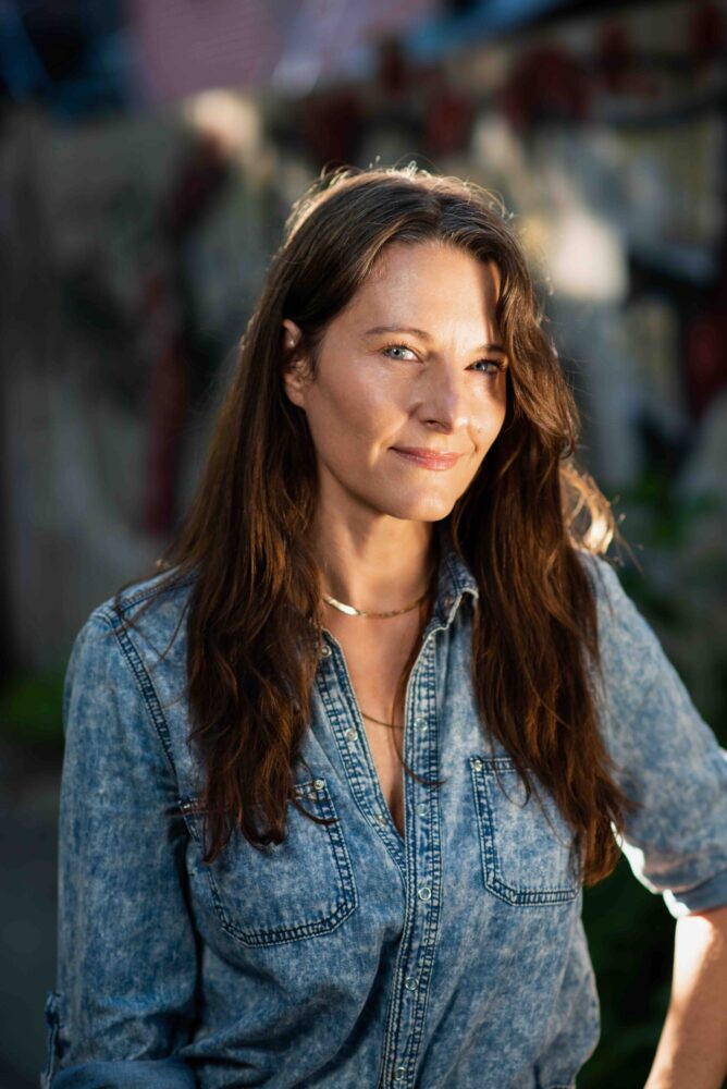 Portrait of Shannon Walsh, wearing a denim shirt and gold necklace, with soft sunlight highlighting her face.