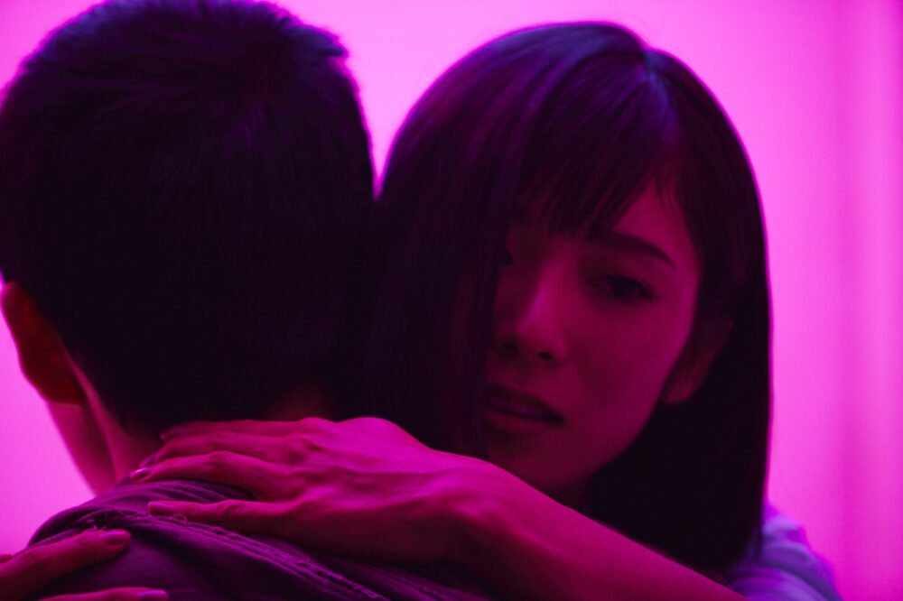 Woman embracing a man in a scene from the film Shoplifters, bathed in pink light.