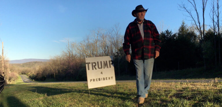 Man stands by a "Trump 4 President" sign in a rural setting. Road in the background.