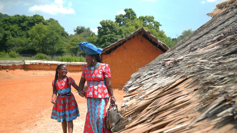 Woman and child in traditional African clothing walking near a thatched roof hut.