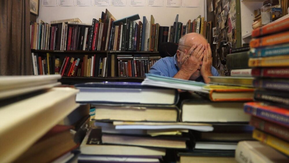 Man overwhelmed by books in a cluttered room covering his face with his hands.