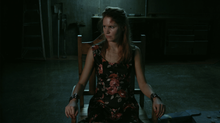 Woman in floral dress, restrained in chair, looks concerned. Dark, suspenseful scene.