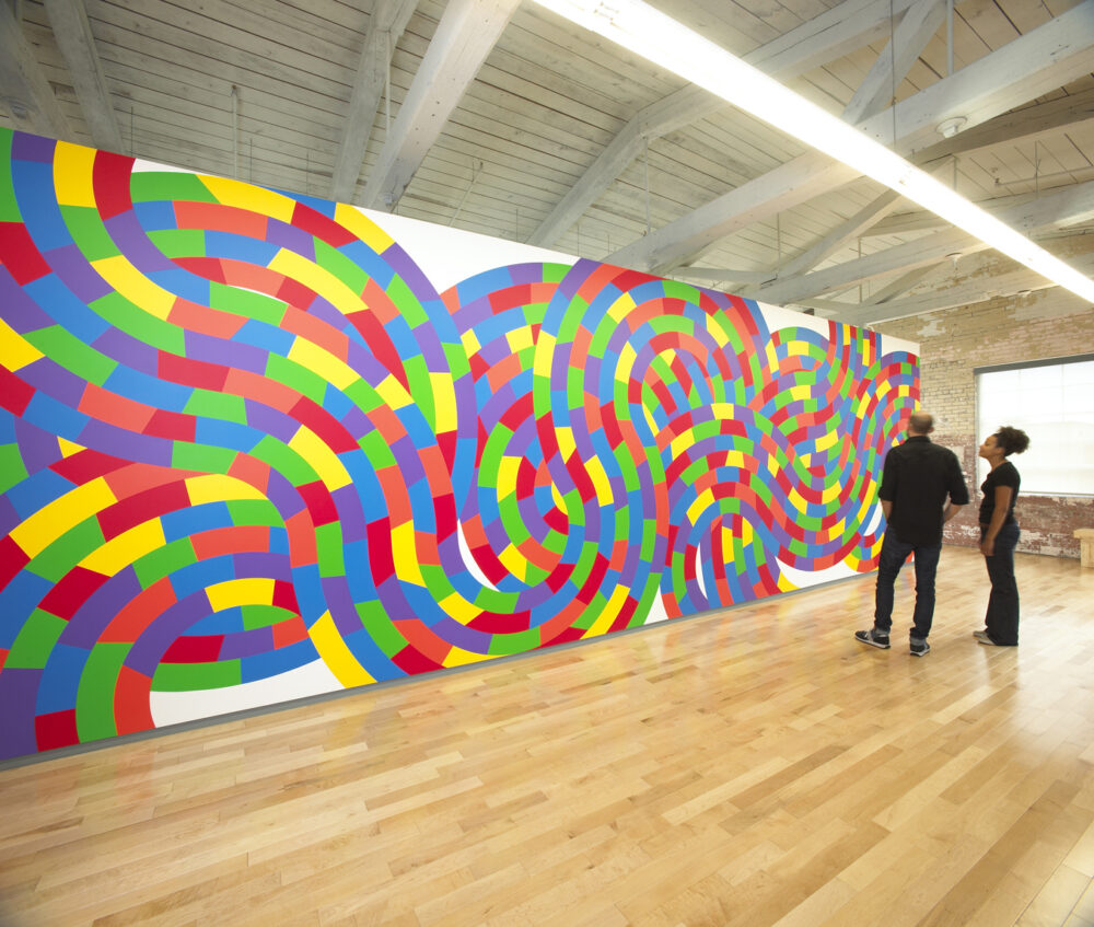 People viewing a vibrant Sol Lewitt wall drawing with colorful geometric patterns in an art gallery.