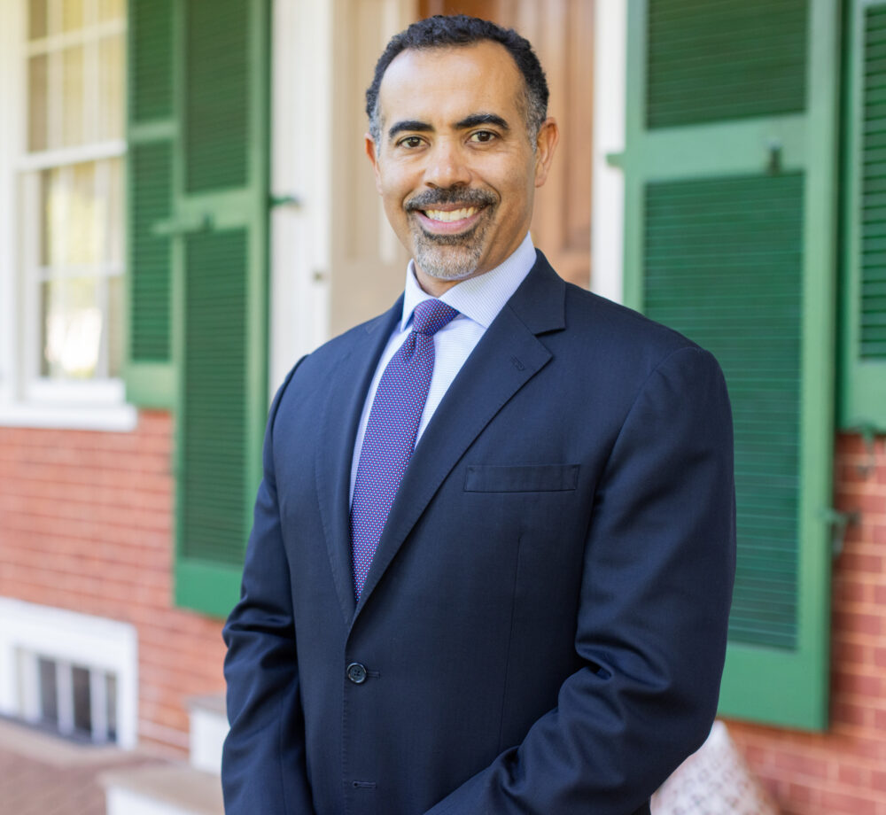 Ian Solomon, smiling in a suit and tie, stands in front of a brick building with green shutters.