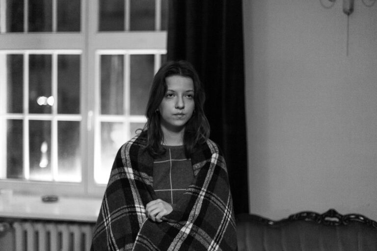 Pensive young woman wrapped in a plaid blanket by a window.