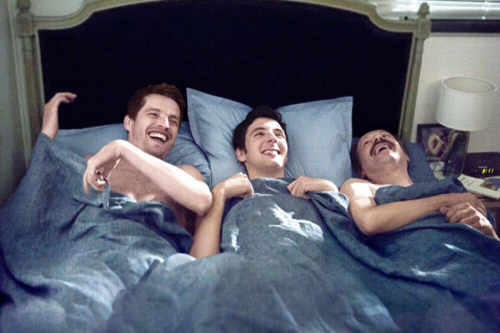 Three men laughing in bed under blue covers.