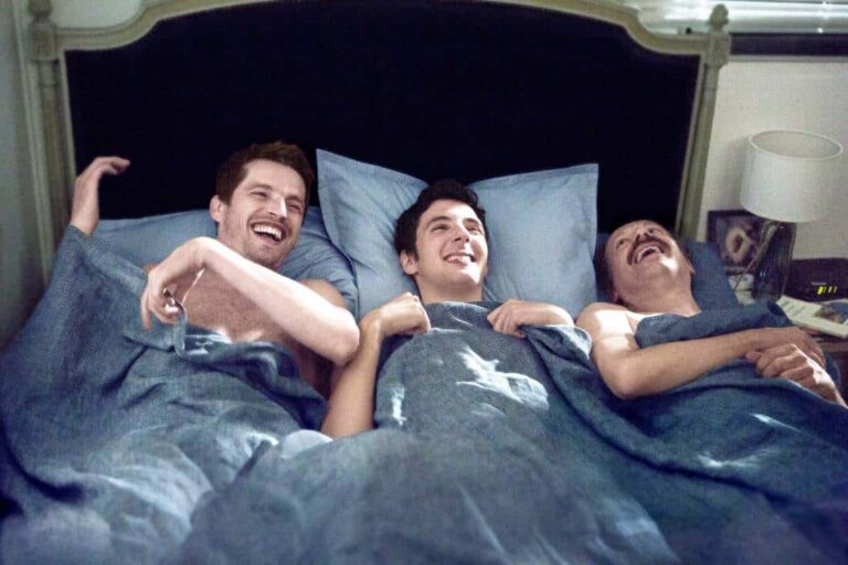 Three men laughing in bed under blue covers.