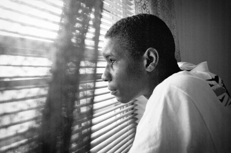 Young man looking out window blinds, deep in thought. Black and white portrait.