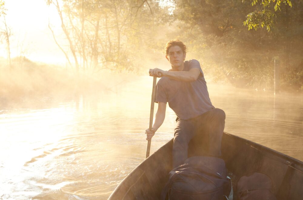 Man in boat, standing and using paddle amidst misty water and trees.