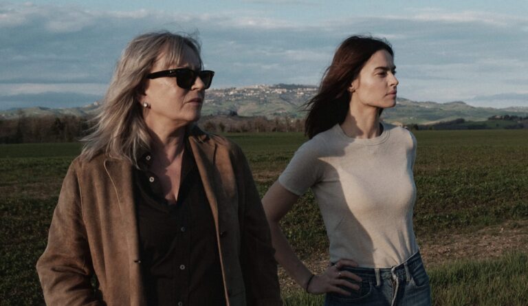 Two women stand in a field overlooking a Tuscan town.