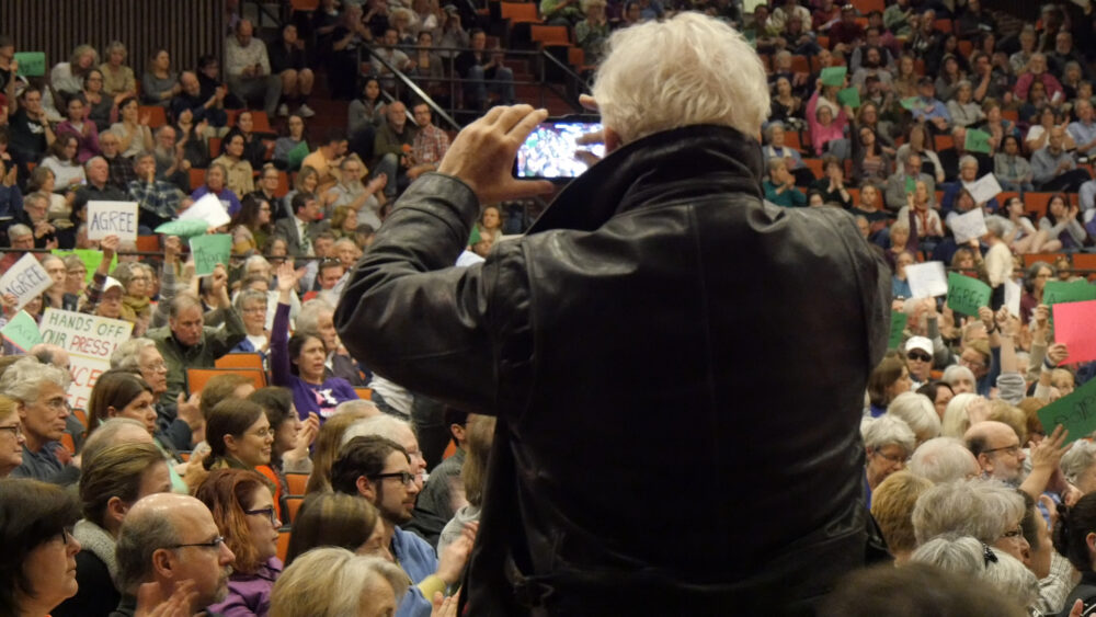 Man in leather jacket photographs a crowd holding signs at an event.