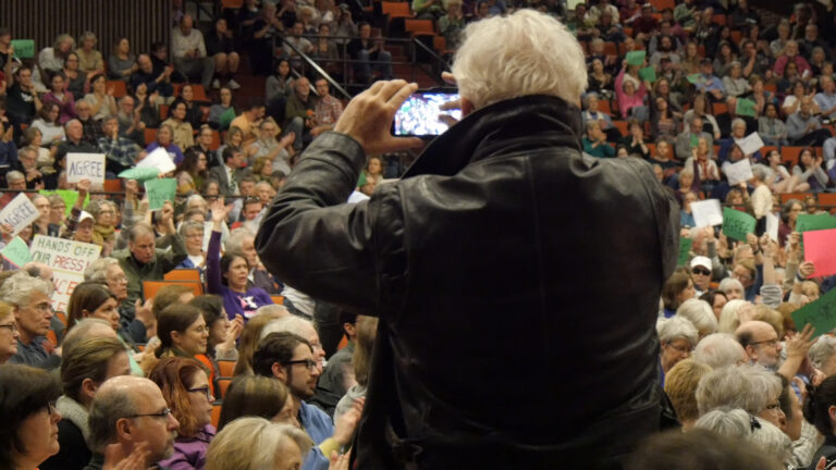 Man in leather jacket photographs a crowd holding signs at an event.