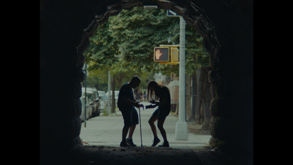 Two people facing each other on a street, seen through a stone archway with a "Don't Walk" sign.