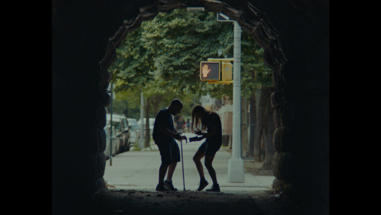 Two people facing each other on a street, seen through a stone archway with a "Don't Walk" sign.