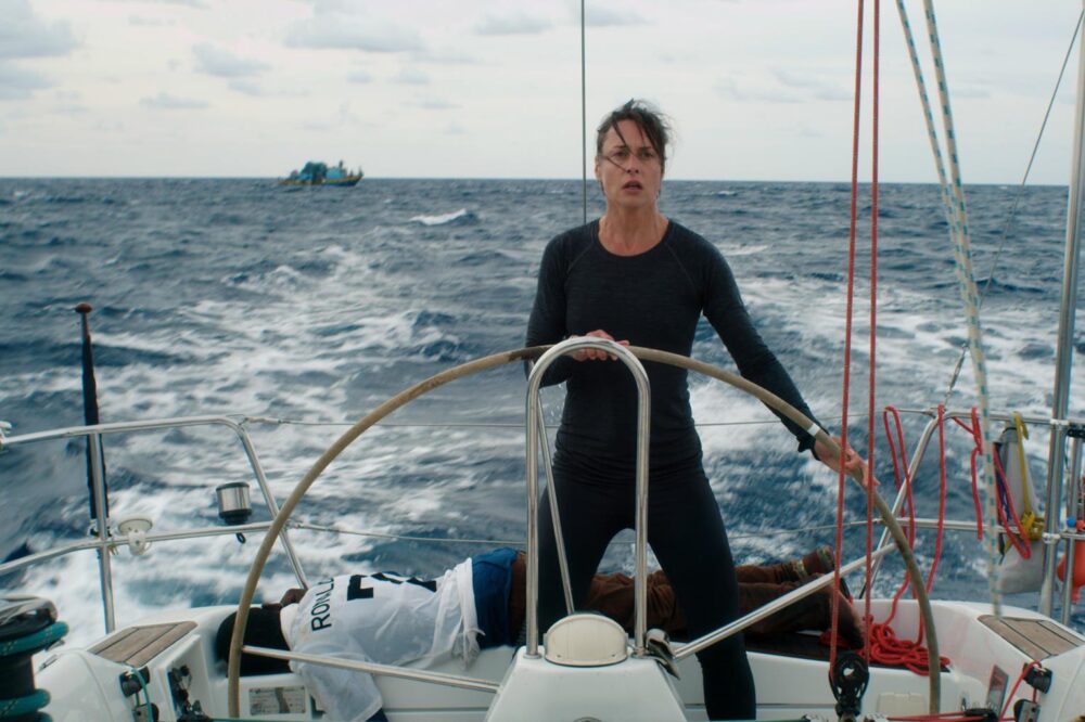 Woman sailing solo on a yacht, possibly related to the film "Styx," with a figure lying on the deck.