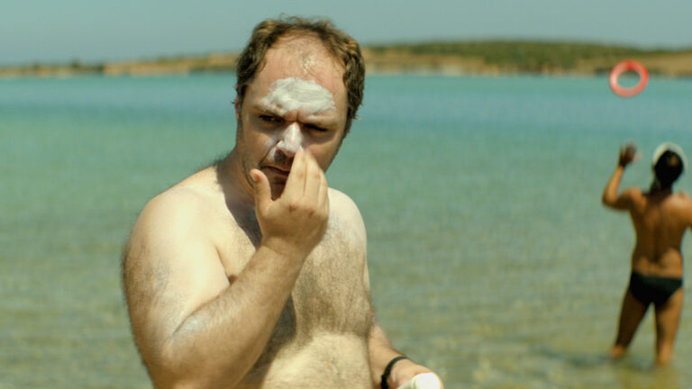 Man applying sunscreen on his face at the beach. A person plays with a ring float in the background.
