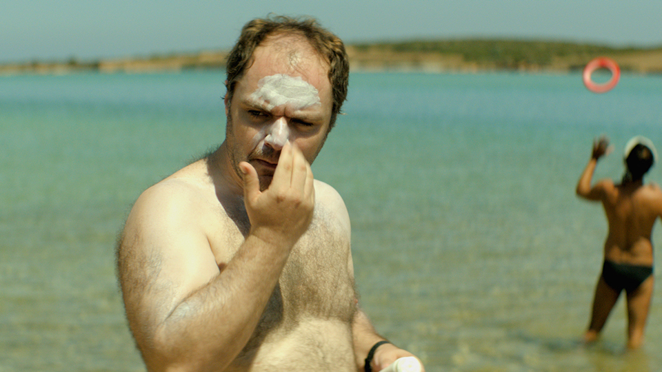 Man applying sunscreen on his face at the beach. A person plays with a ring float in the background.