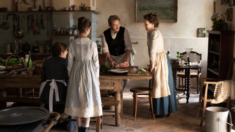 Scene from "The Taste of Things" movie: a chef preparing food while women watch in a rustic kitchen.