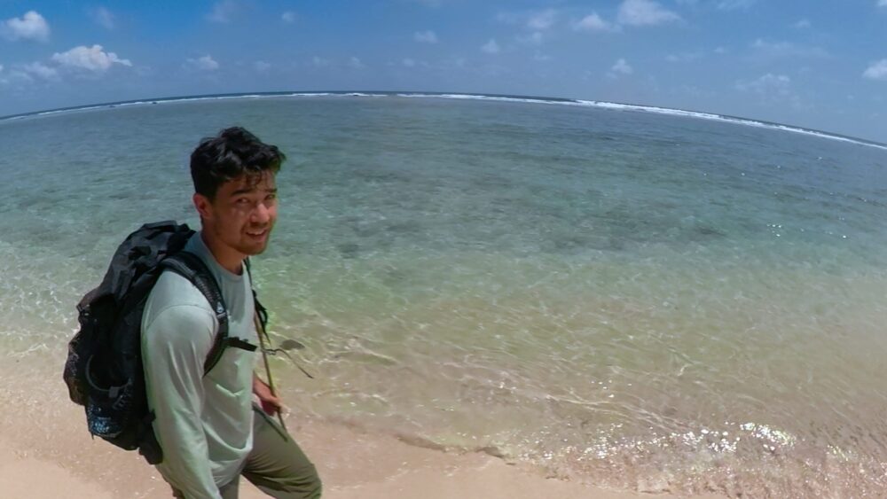 Man with backpack walks along a tropical beach with clear water and a blue sky.