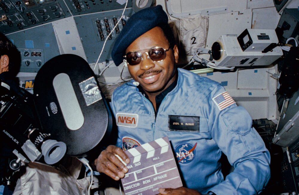 Astronaut Ronald McNair in space with a camera, holding a clapperboard for "American Adventure.
