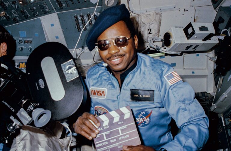 Astronaut Ronald McNair in space with a camera, holding a clapperboard for "American Adventure.
