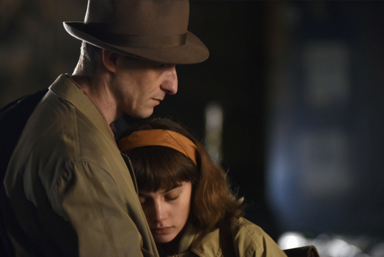 Scene from "Those Who Remained" film: a man in a hat embraces a young woman with a headband, both looking somber.