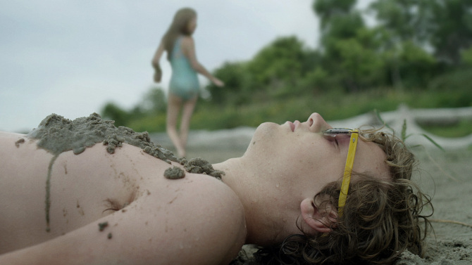 Boy covered in sand lying on a beach, girl in swimsuit in background.