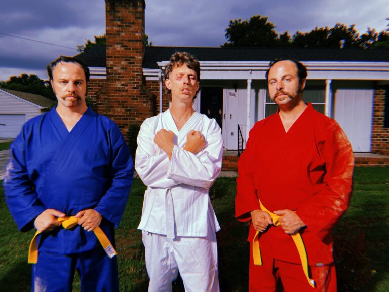 Three men in karate uniforms with yellow belts pose in front of a house.