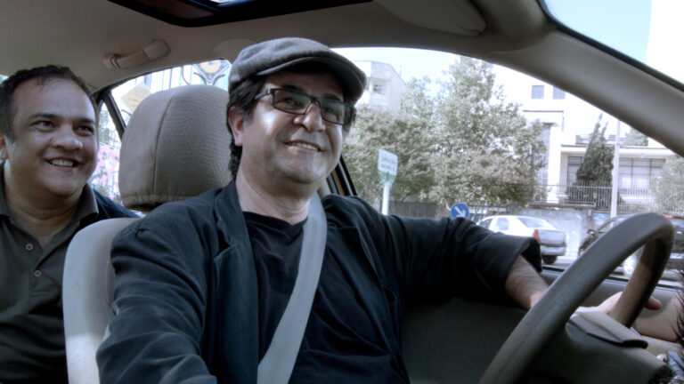 Man driving a car with a smiling passenger in the back seat, wearing a cap and glasses.