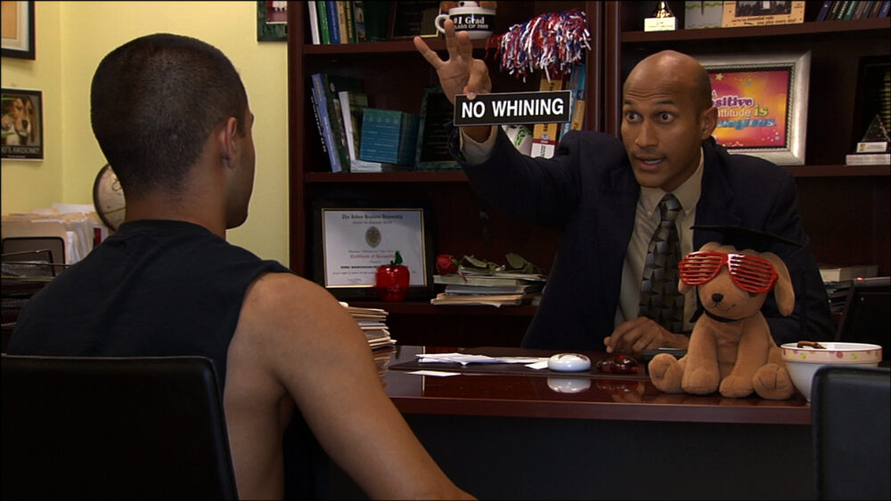 Teacher sitting at a desk holding "No Whining" sign while talking to student.