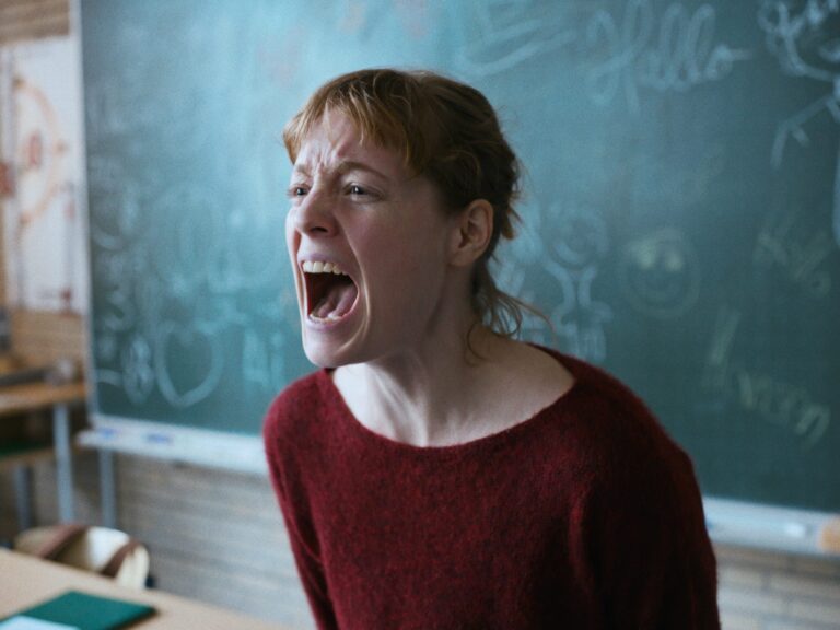 Frustrated teacher in a red sweater yells in front of a chalkboard. Classroom scene.