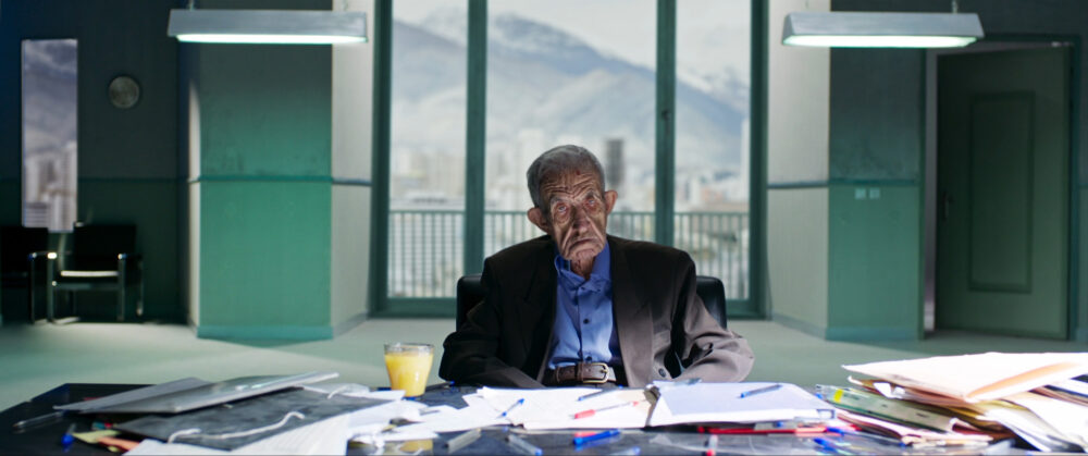 Elderly man sits at a desk with papers, pens, and juice, with a mountain view backdrop.
