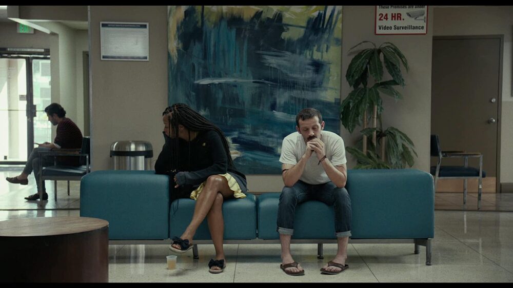 Two people sit in a waiting room with a "Video Surveillance" sign.