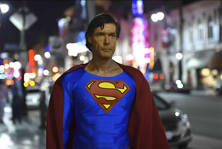 Superman stands on a city street at night, cape blowing in the wind.