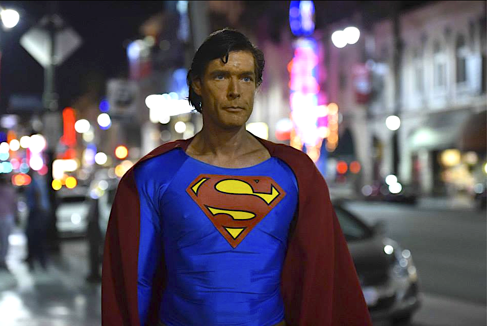Superman stands on a city street at night, cape blowing in the wind.