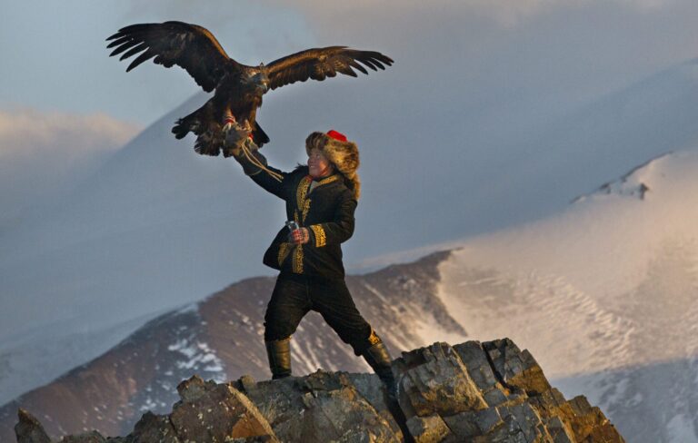 Eagle huntress with golden eagle perched on her arm, standing on a rocky mountain peak.
