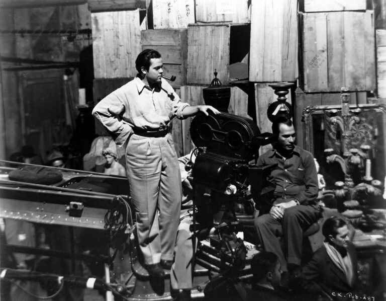 Orson Welles on set, smoking a pipe, with film crew and equipment.