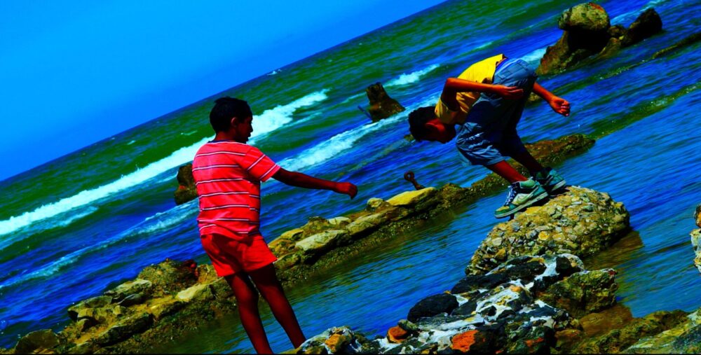 Boys playing on rocks by the ocean. One boy reaches out to help another climbing.