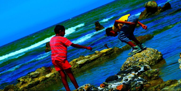 Boys playing on rocks by the ocean. One boy reaches out to help another climbing.