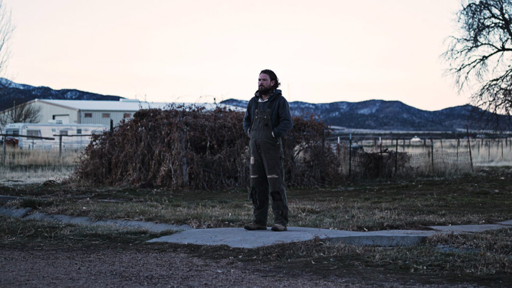 Man in overalls stands alone in a desolate landscape, mountains in background.