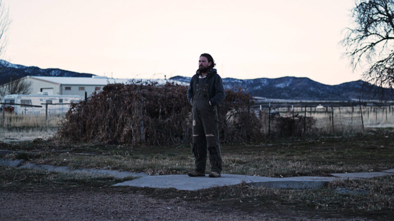 Man in overalls stands alone in a desolate landscape, mountains in background.