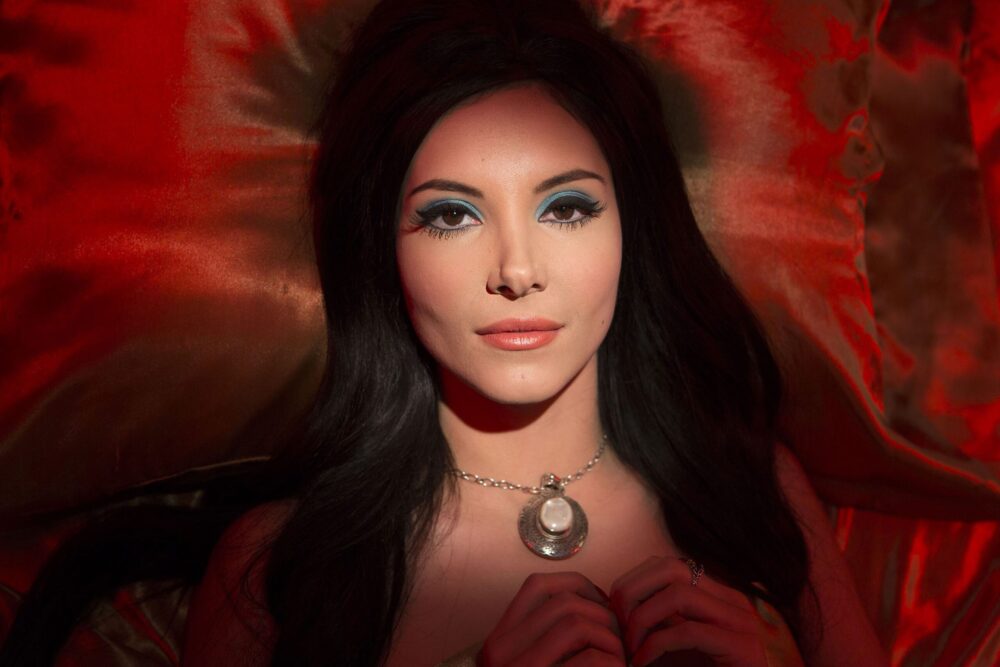 Woman with vintage makeup and a pendant laying on a red silk pillow.