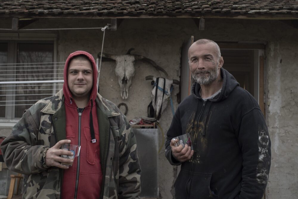 Two men holding drinks in front of a rural house with animal skulls.