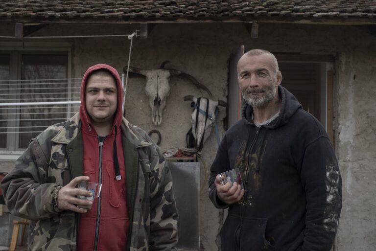Two men holding drinks in front of a rural house with animal skulls.