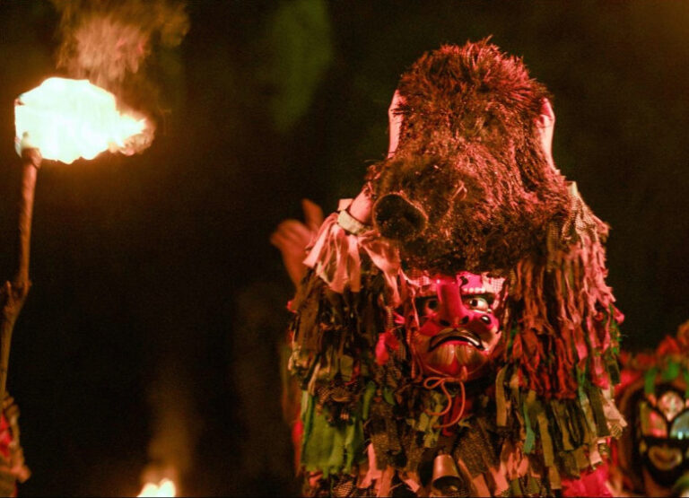 Costumed figure in a red mask and fringed garment holds a torch.