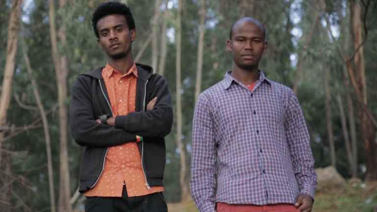 Two young men stand in a wooded area. One wears a black hoodie, the other a plaid shirt.