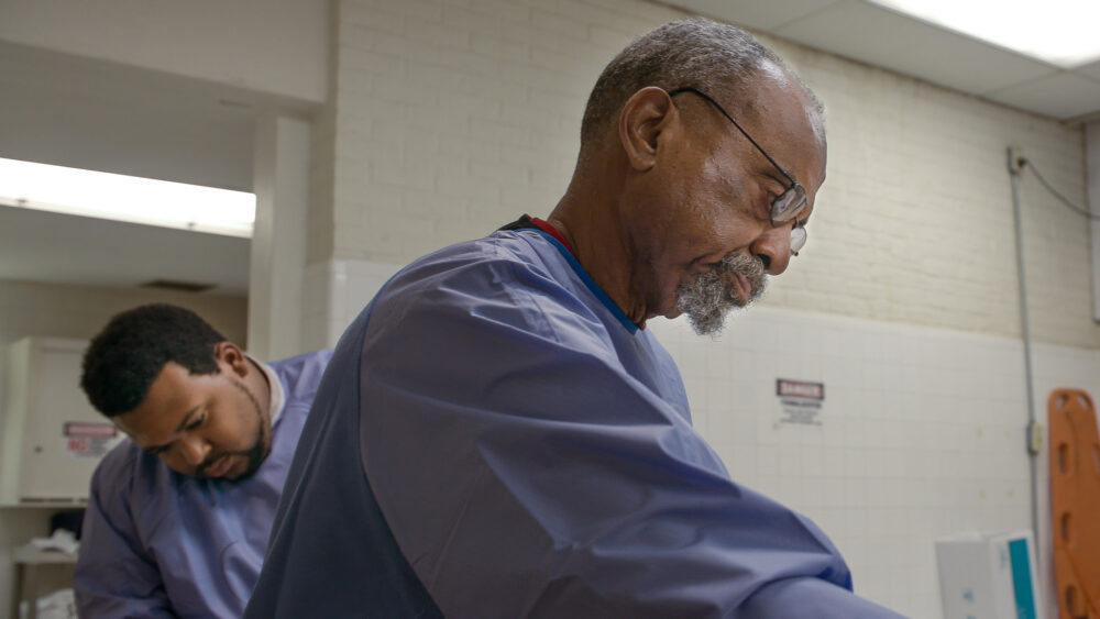 Two medical examiners in scrubs working in a morgue, one with gray hair and glasses.